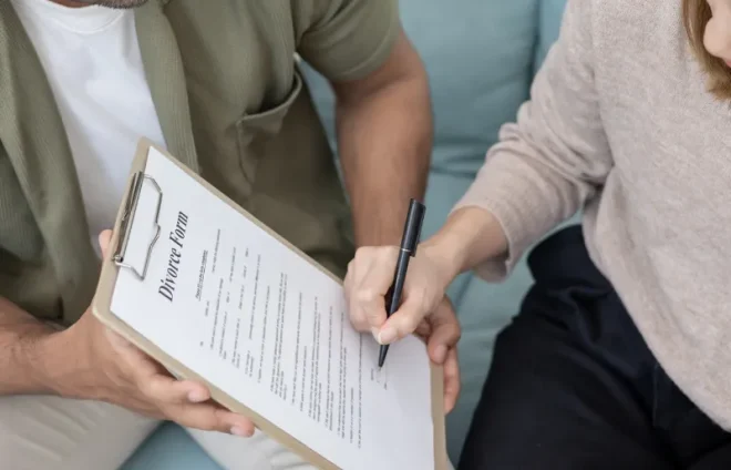 Couple reviews clipboard document while spouse signs divorce form together on living room sofa calmly