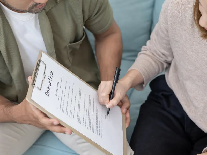 Couple reviews clipboard document while spouse signs divorce form together on living room sofa calmly