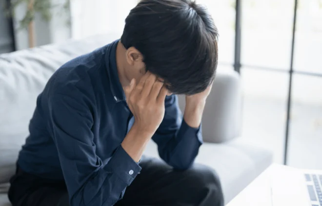 Worried man holds head while sitting near laptop, feeling overwhelmed by bills and finances today