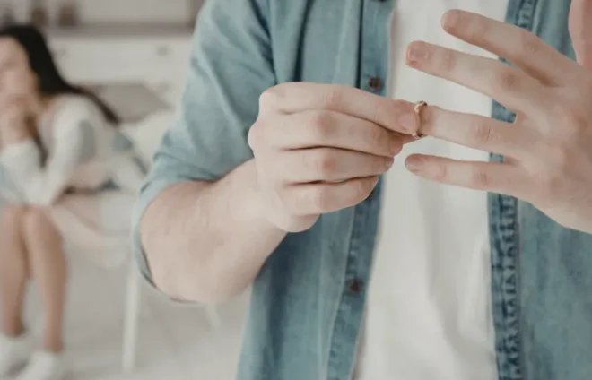 Man removes wedding ring, considering divorce, while upset partner sits blurred in background at home