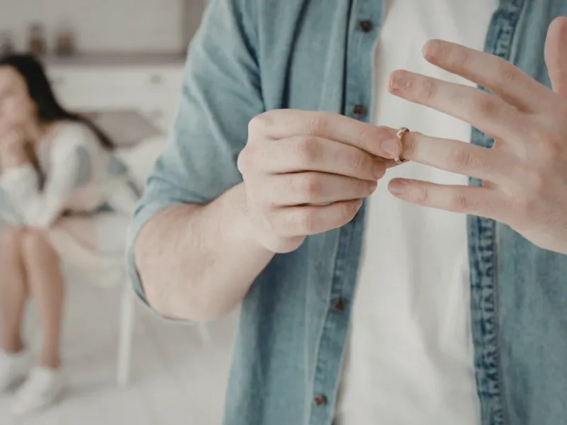 Man removes wedding ring, considering divorce, while upset partner sits blurred in background at home