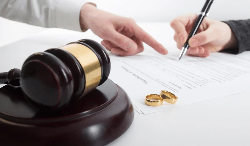 Gavel and wedding rings beside document as person signs divorce agreement under legal guidance today