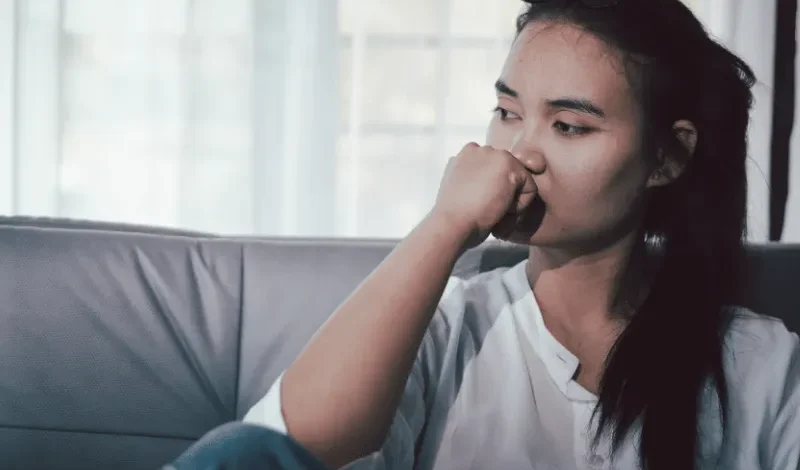 Woman sitting on a couch with her hand near her mouth, looking away in a quiet indoor setting