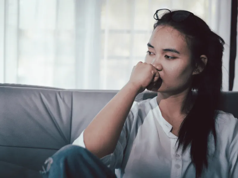 Woman sitting on a couch with her hand near her mouth, looking away in a quiet indoor setting