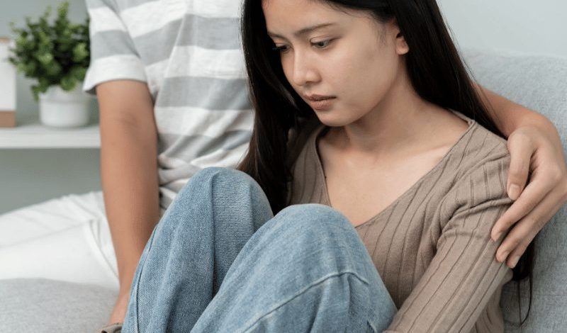 Worried woman sitting on sofa while partner comforts her, showing emotional strain during financial hardship