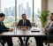 Three professionals seated around a meeting table in a high-rise office with large windows and a city view