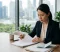 Woman in business attire reviewing documents at a desk in a bright office with a city skyline behind her