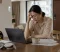 Workplace Stress Divorce A tired professional sits at a dining table with a laptop, documents, coffee, and family items, showing how workplace stress may lead someone to seek advice from a divorce lawyer