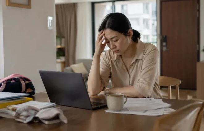 A tired professional sits at a dining table with a laptop, documents, coffee, and family items, showing how workplace stress may lead someone to seek advice from a divorce lawyer