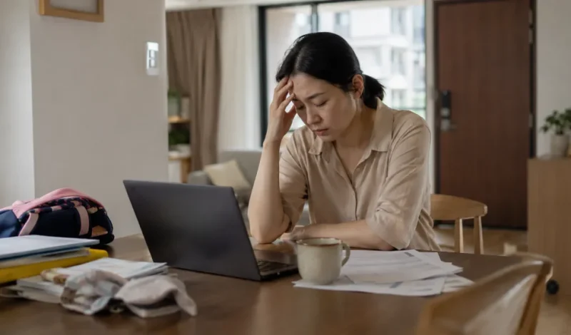 A tired professional sits at a dining table with a laptop, documents, coffee, and family items, showing how workplace stress may lead someone to seek advice from a divorce lawyer